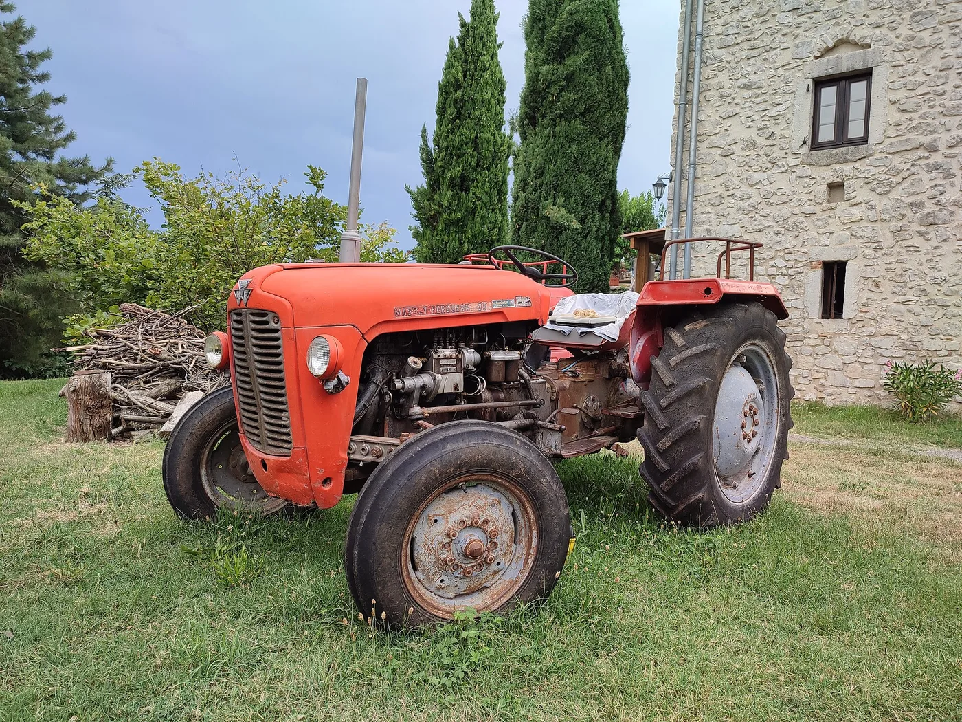 Tracteur Massey Ferguson 35 rouge et gris, moteur apparent, garé dans une cour en herbe devant une maison en pierre